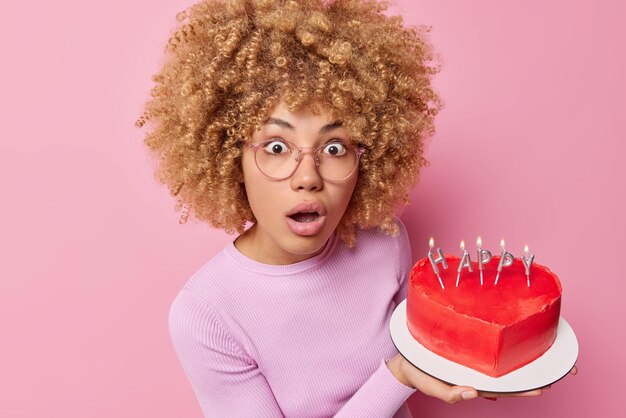 巻き毛のふさふさした髪の驚きのショックを受けた女性は、ハート型のおいしいケーキを持って誕生日パーティーにあなたを招待しますピンクの壁に隔離された眼鏡カジュアルジャンパーを着ています