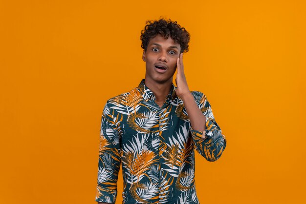 Surprised and shocked young handsome dark-skinned man with curly hair in leaves printed shirt keeping hand on head on an orange background