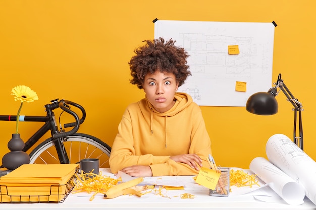 Free photo surprised skilled female engineer works on building blueprint stares shocked, realises deadline involved in working process dressed in sweatshirt