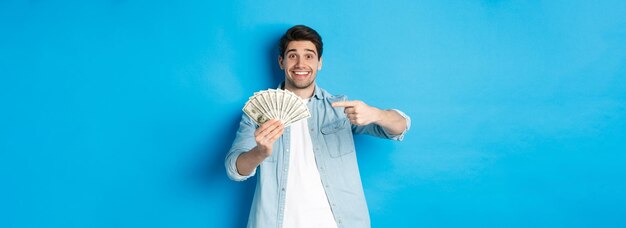 Foto gratuita uomo sorridente sorpreso in abiti casual che punta il dito contro i soldi in piedi su sfondo blu