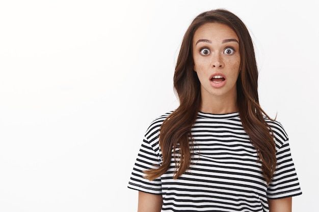 Surprised speechless cute brunette girl in striped t-shirt drop jaw from amazement, stare camera see amazing promo, gasping astonished popping eyes impressed, standing white wall