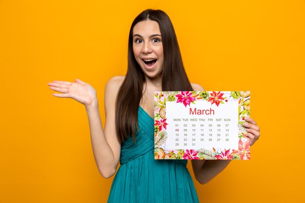 Удивленная протягивающая руку красивая молодая девушка в счастливый женский день держит календарь