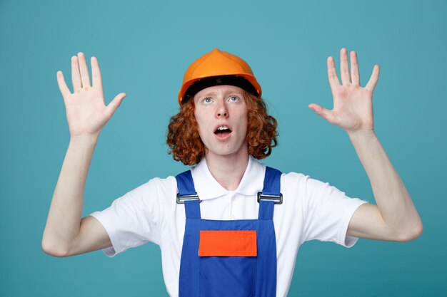 surprised spreading hands young builder man in uniform isolated on blue background