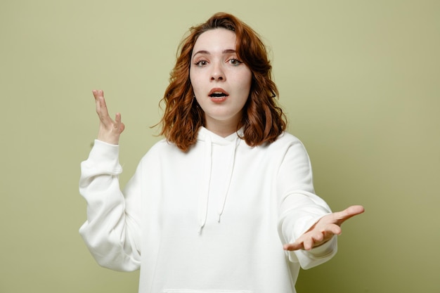 Удивленно разводя руками молодая женщина в белом свитере на зеленом фоне