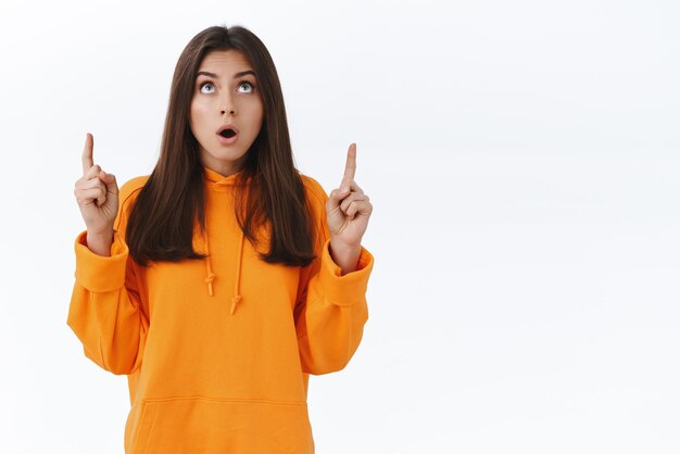 Surprised startled cute brunette female student found something cool upwards open mouth say wow and pointing fingers up at special offer advertisement with awesome discounts white background