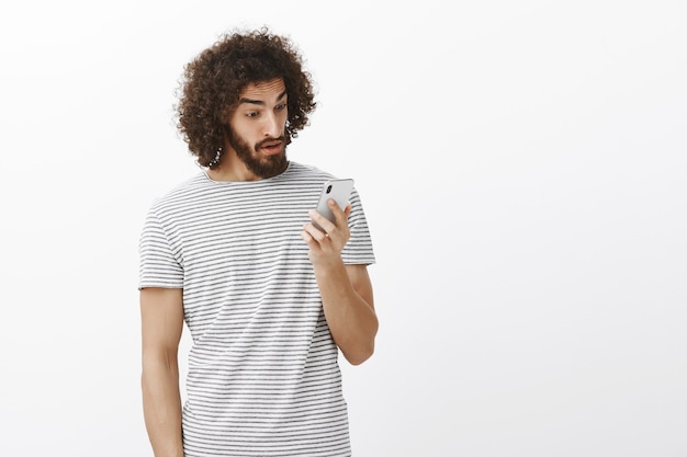 Surprised stunned emotive guy with curly hair and beard, staring at smartphone screen with amazed expression