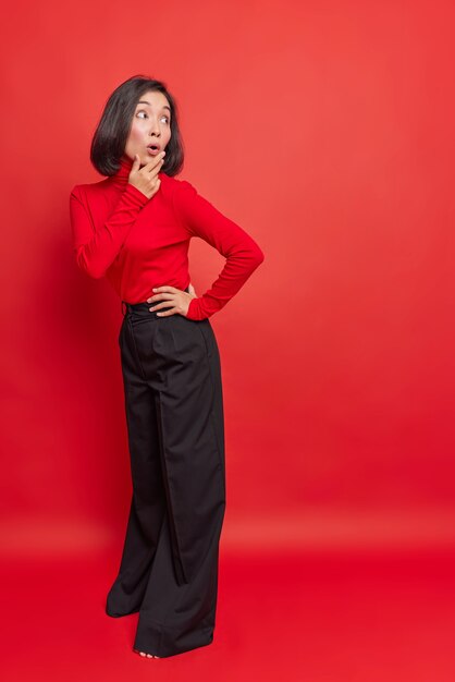 Surprised stylish dark haired Asian woman looks back with shocked expression wears turtleneck and loose black trousers poses against vivid red wall copy space for your promotional content