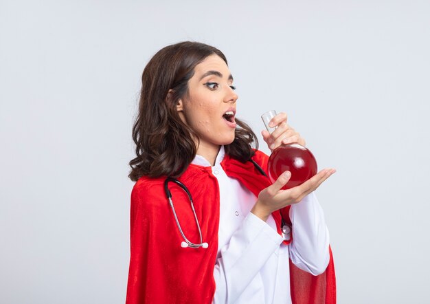 Surprised superwoman in doctor uniform with red cape and stethoscope holding and looking at red chemical liquid in glass flask isolated on white wall