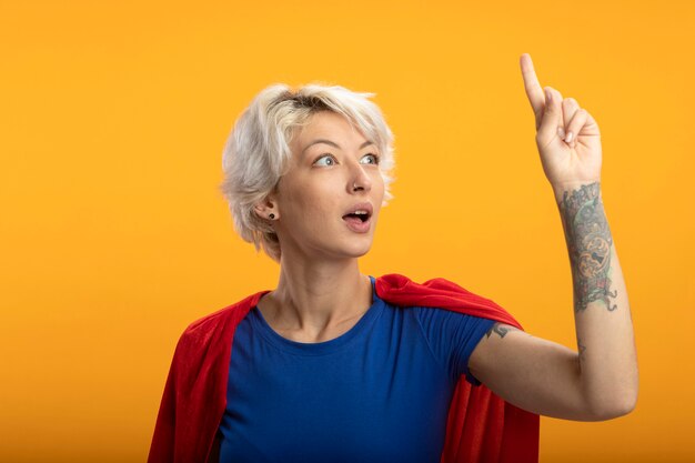 Free photo surprised superwoman with red cape points up isolated on orange wall