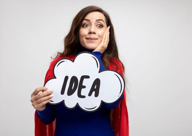Surprised superwoman with red cape puts hand on face and holds idea bubble looking at side isolated on white wall