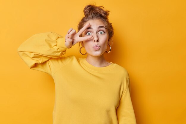 Surprised teenage girl keeps lips folded makes peace gesture with fingers over eye shows salute sign wears big round silver earrings and jumper isolated over yellow background. Body language concept