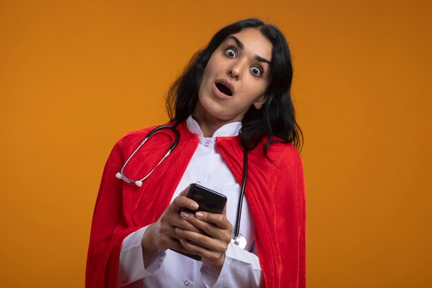 Surprised tilting head young superhero girl wearing medical robe with stethoscope holding phone isolated on orange wall