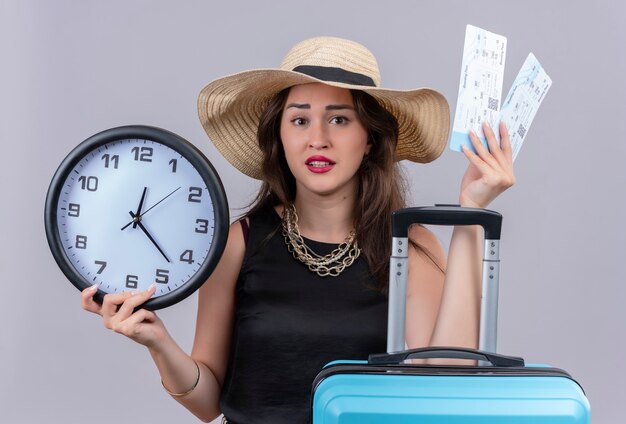 Surprised traveler young girl wearing black undershirt in hat holding suitcase and tickets on white background