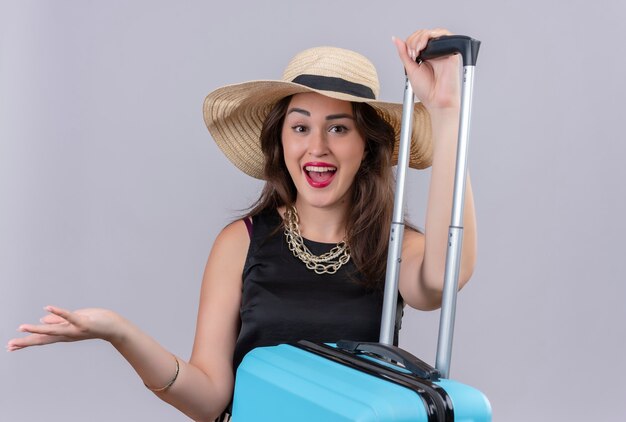 帽子に黒のアンダーシャツを着て驚いた旅行者の若い女の子は、白い背景の上のスーツケースに手を置いた