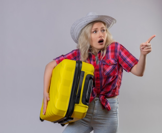 Surprised traveler young girl wearing red shirt in hat holding suitcase points to side on isolated white background