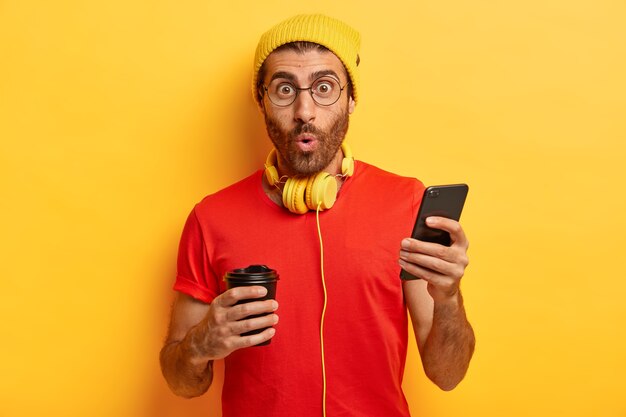 Surprised unshaven guy listens music in headphones, sends text messages on cell phone, types answer