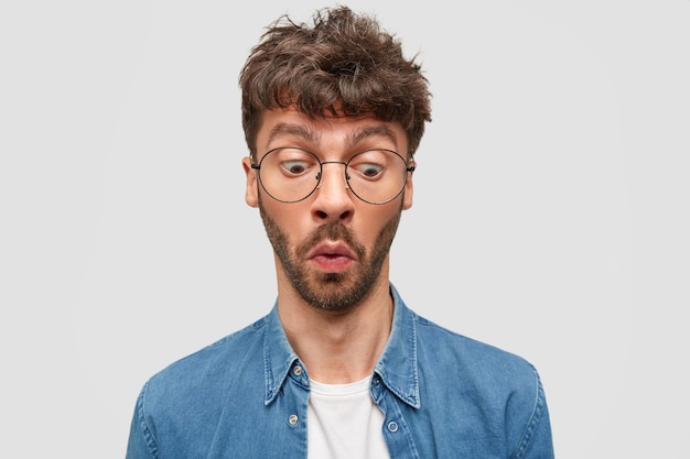 Surprised unshaven man wonders while notices something down, has displeased expression, wears glasses and shirt