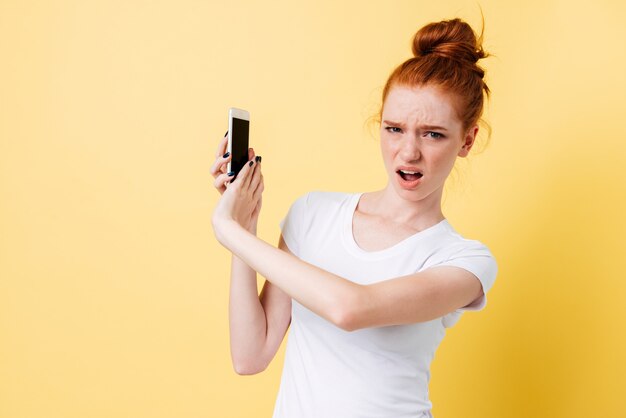 Surprised upset ginger woman in t-shirt covering smartphone in hands