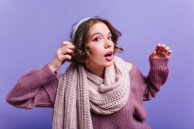 Surprised white girl in long knitted scarf posing on purple wall. emotional brunette lady in woolen accessories.