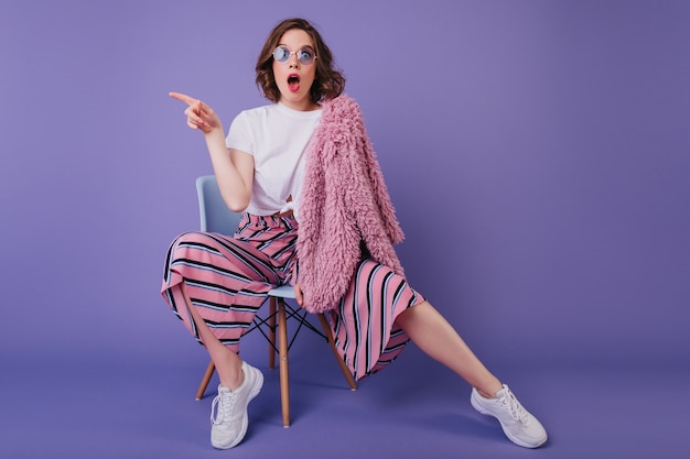 Surprised white woman in striped pants sitting on purple wall with mouth open. Indoor photo of amazed curly girl in fur jacket posing on chair .