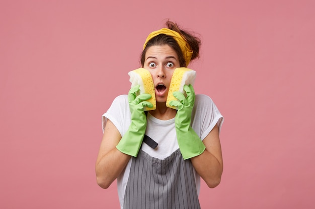 Удивленная женщина в фартуке и повседневной одежде в зеленых резиновых перчатках держит на щеках две аккуратные губки, понимая, что ей предстоит много работать. Удивленная женщина собирается делать свою работу по дому