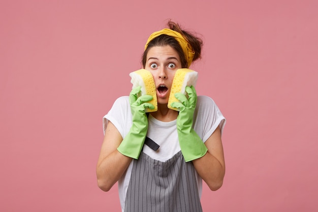 Surprised woman in apron and casual clothes wearing rubber green gloves holding two tidy sponges on cheeks realising that she should do much work. Astonished female going to do her housework