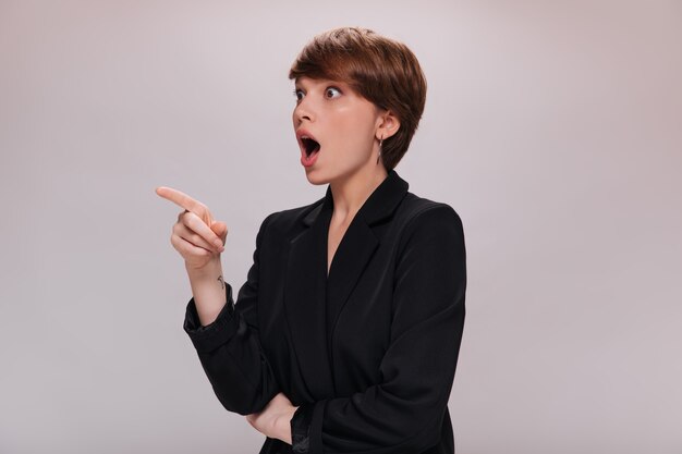 Surprised woman in black suit points to left. Attractive dark-haired lady in classic-style jacket looks shocked on isolated background