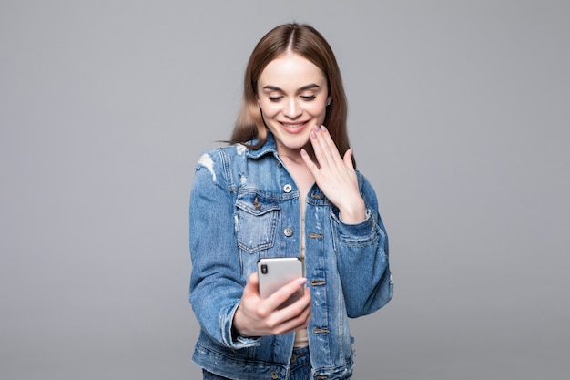 Surprised woman covering mouth, looking at mobile phone screen, shocked female reading unexpected message, shopping offer, good news, holding cellphone, isolated on grey wall