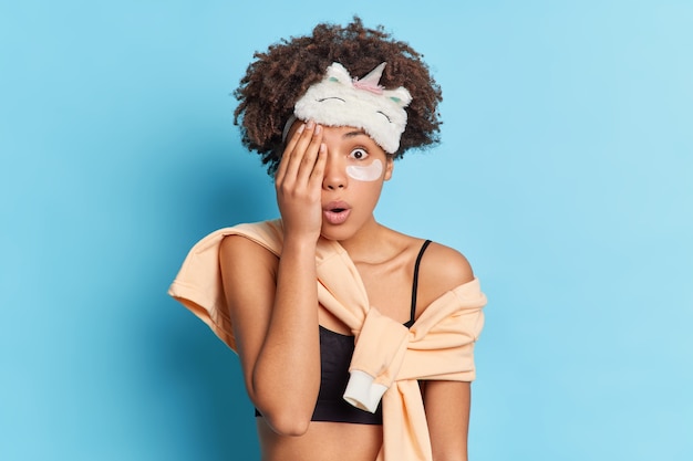 Free photo surprised woman covers eye with hand stares at camera hides face applies collagen patches take care of her skin dressed in pajama wears sleepmask on forehead isolated on blue wall