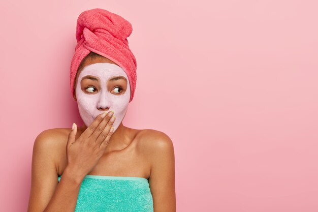 Surprised woman covers mouth, applies nourishing mask for removing dead cells, wears wrapped towel on head, stands against pink wall