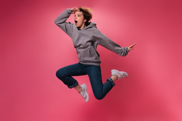 Foto gratuita donna sorpresa in felpa con cappuccio grigia che salta su sfondo rosa. ragazza dai capelli corti scioccata in jeans si muove e balla su sfondo isolato