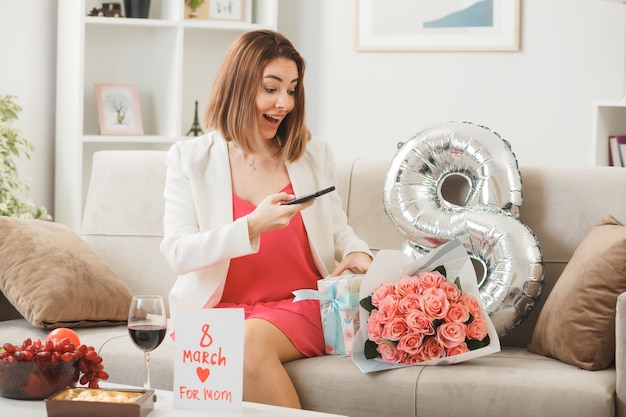 Удивленная женщина в счастливый женский день держит телефон, сидя на диване в гостиной