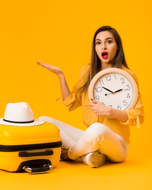 Surprised woman holding clock next to luggage with hat on top