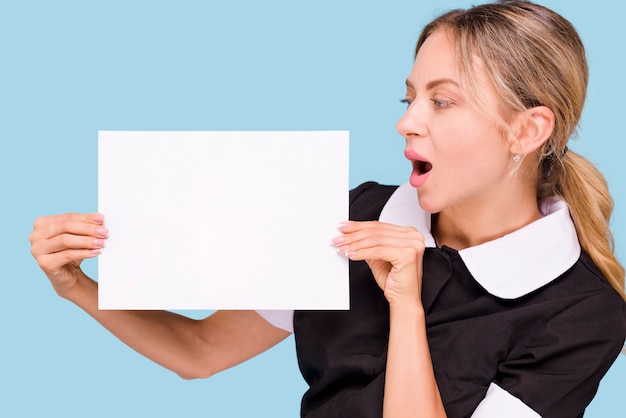 Surprised woman holding and looking at blank white paper standing against blue wall