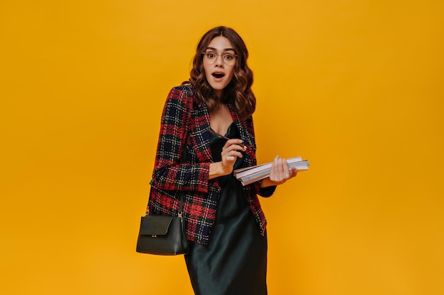 Surprised woman holds books Beautiful curly lady in checkered jacket and dress looks into camera on orange background