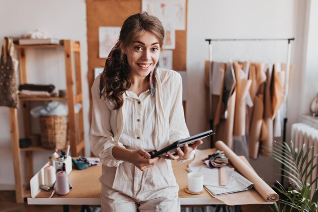 Surprised woman in light suit holds computer tablet and leans on table Happy longhaired curly girl in beige shirt smiling in designer office