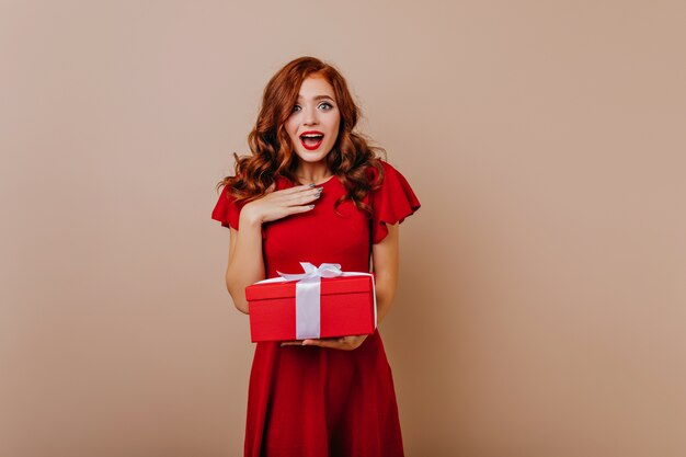 Surprised woman in red dress holding presents. Attractive ginger girl celebrating birthday.