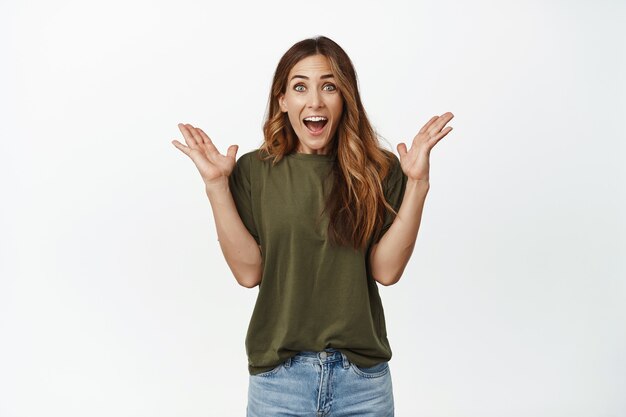 Surprised woman screaming and looking excited at front, standing against white wall