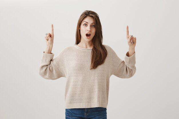 Surprised woman showing promo, pointing fingers up and drop jaw shocked