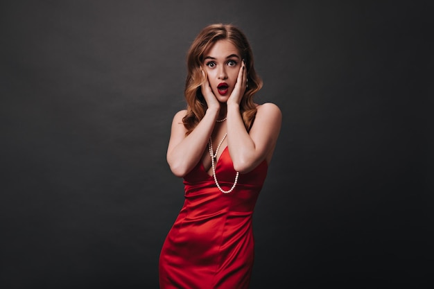 Surprised woman in silk dress and pearl jewelry posing on black background Curly lady in red gorgeous outfit looks shocked