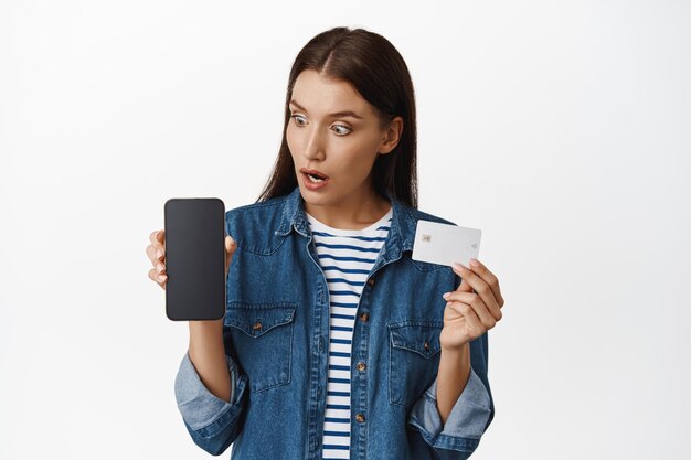 Surprised woman stare at her phone, showing smartphone screen and credit card, amazing prices online, shopping in internet via mobile app, white background. Copy space