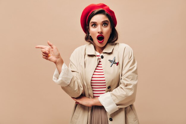 Surprised woman in trench coat pointing to place for text on beige background. Modern girl in long coat and hat posing and looking into camera.