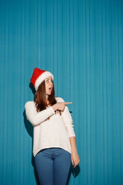 파란색 배경에 오른쪽을 가리키는 산타 모자를 쓰고 놀란 된 여자