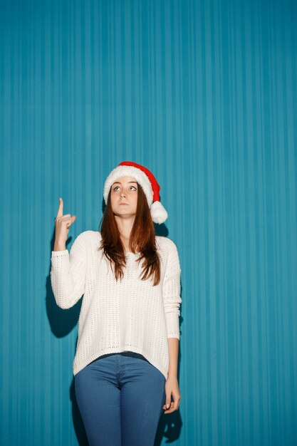 surprised woman wearing a santa hat pointing up on blue studio background