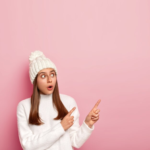 Surprised woman wears white hat and sweater, has startled look aside, drops jaw, demonstares something strange, poses against pink wall
