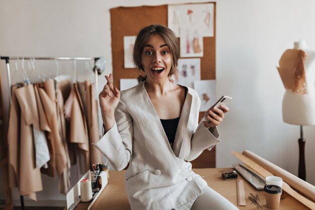 Surprised woman in white suit crosses fingers and holding phone