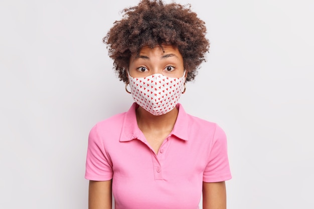 Surprised woman with curly hair wears disposable mask during quarantine and coronavirus outbreak cares about health wears casual pink t shirt isolated over white wall