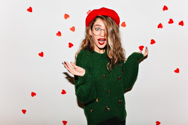 Surprised woman with light-brown hair posing on white wall in green sweater