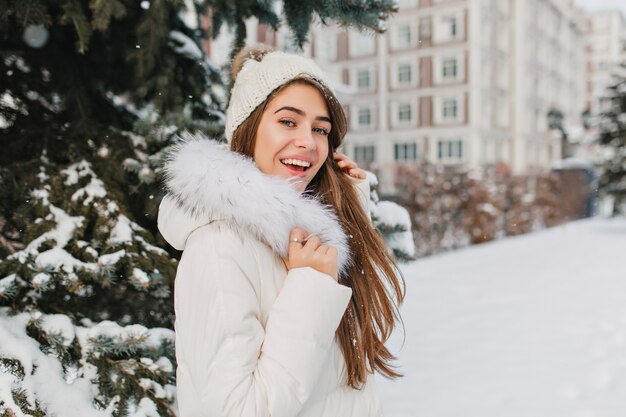 Удивленная женщина с длинными прямыми волосами веселится в зимние каникулы, проводя время на открытом воздухе. Портрет восторженной кавказской женщины в белом наряде охлаждает в парке в снежный день.