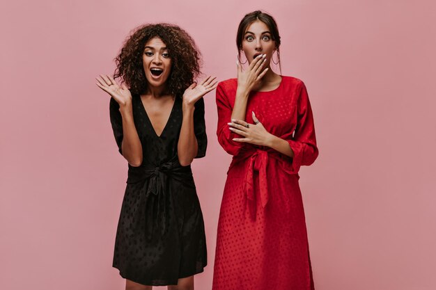 Удивленная женщина с круглыми серьгами в красном платье в горошек смотрит в камеру и позирует с девушкой-мулаткой в черном модном наряде